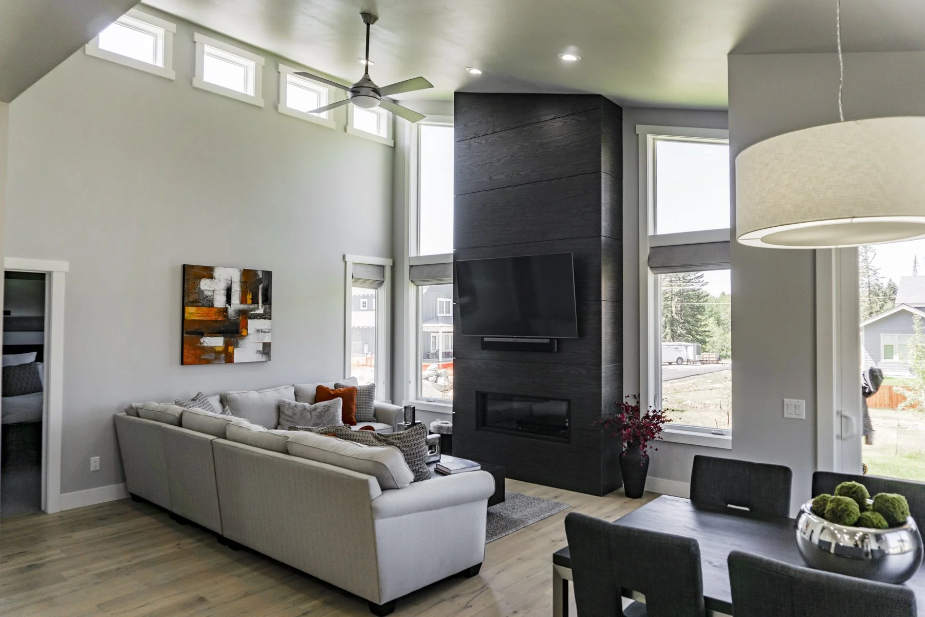 Modern living room with high ceiling, large windows, a beige sectional sofa with throw pillows, black wall with a mounted flat-screen TV, a fireplace, and a ceiling fan.