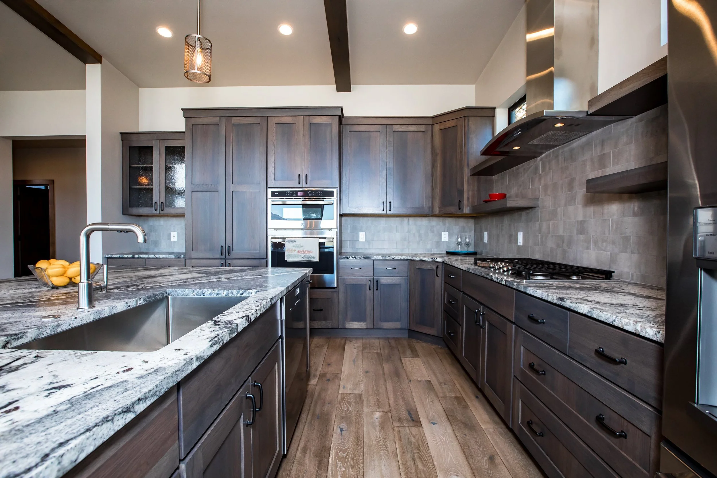Modern kitchen with dark wooden cabinetry, granite countertops, and stainless steel appliances, including double ovens and a range hood. Features a sink with a brushed metal faucet, a bowl of lemons, and wood flooring.