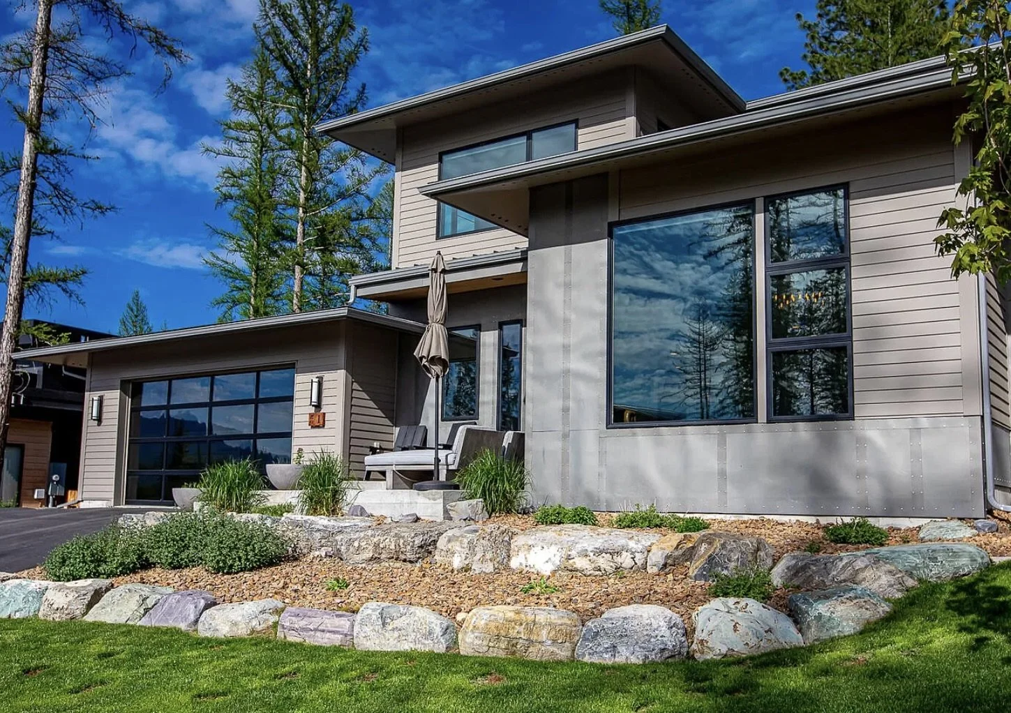 Modern multi-story house with large windows, surrounded by green trees and landscaping with rocks and grass, under a partly cloudy sky.