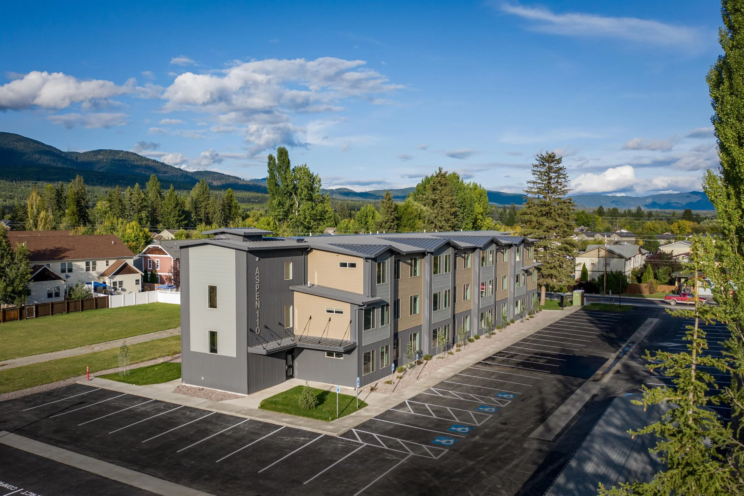 A modern, multi-story apartment building with a parking lot, surrounded by trees and mountains under a partly cloudy sky.