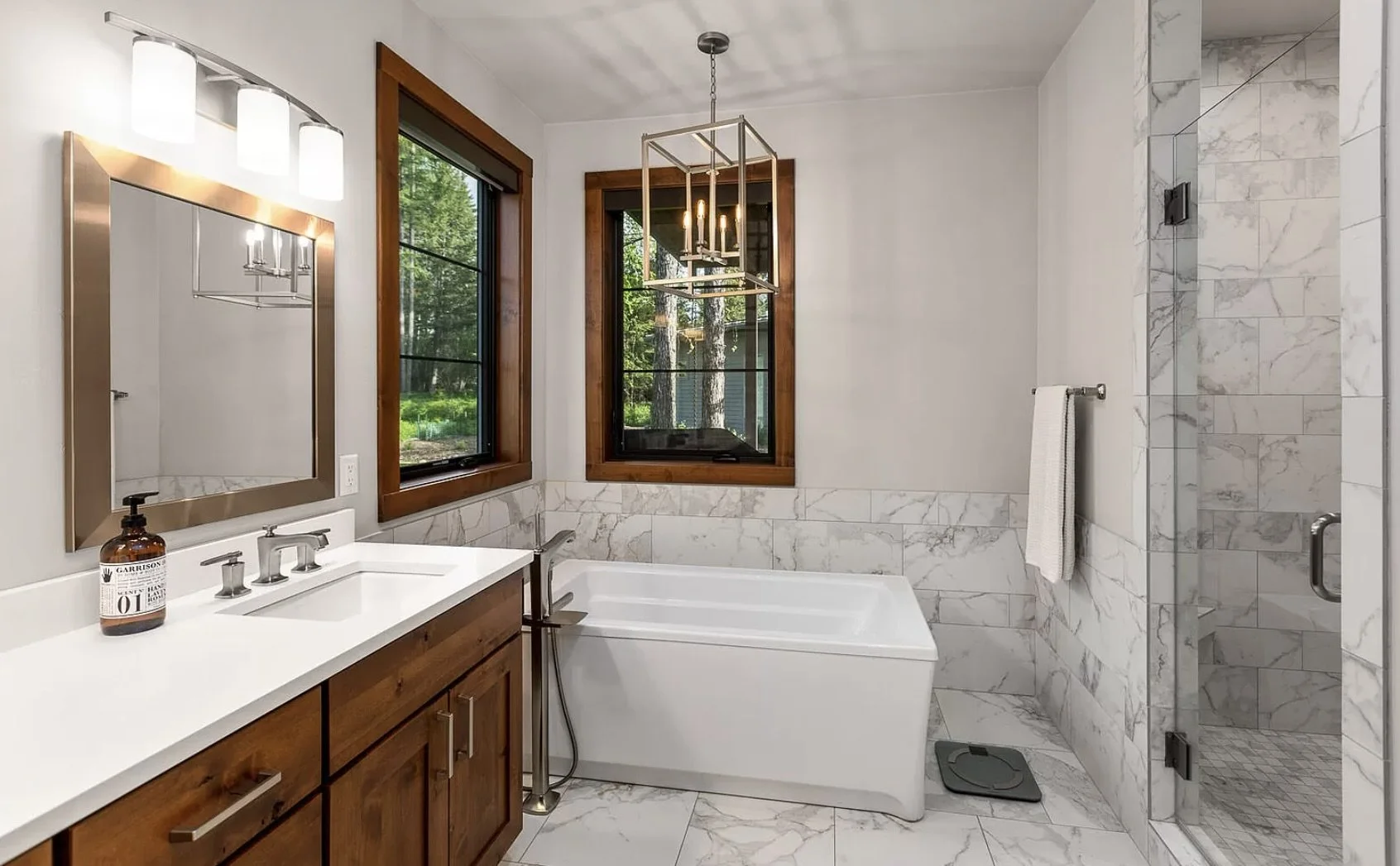 Modern bathroom with white marble tiles, wooden vanity, bathtub, walk-in shower with glass door, and two black-framed windows showing greenery outside.