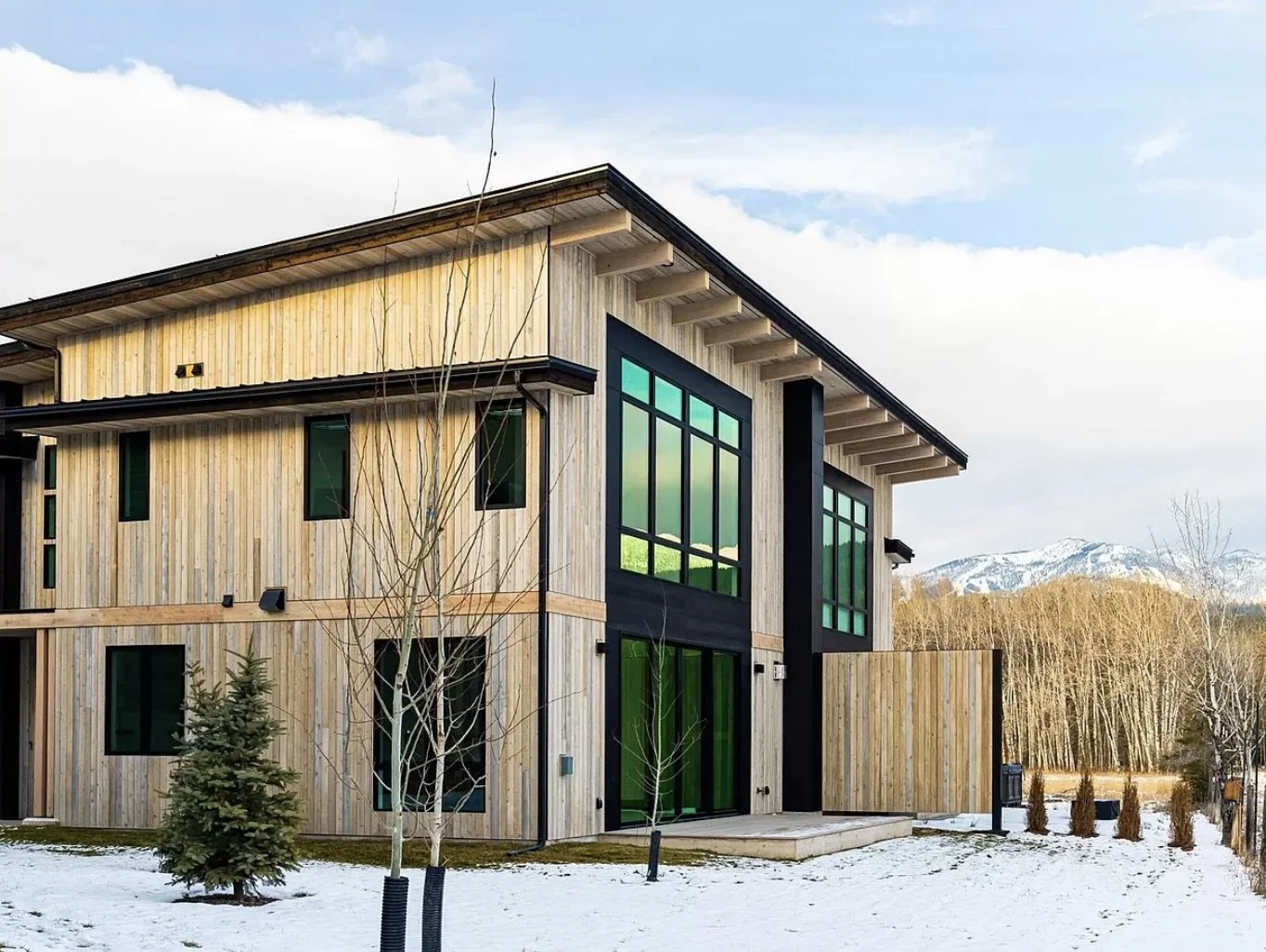 Modern two-story house with wooden exterior, large green windows, and a snow-covered yard, mountains in the background.