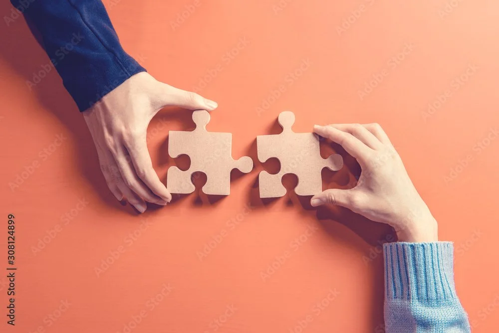 Two hands holding separate puzzle pieces about to connect against a peach-colored background.