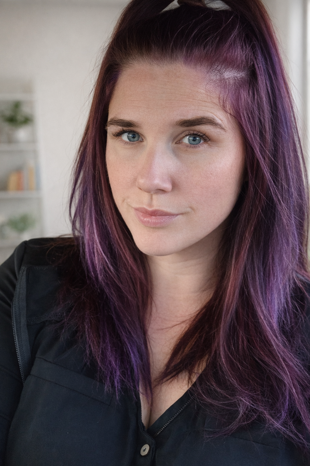 Close-up of a woman with purple hair, fair skin, and blue eyes, wearing a black top, in an indoor setting.
