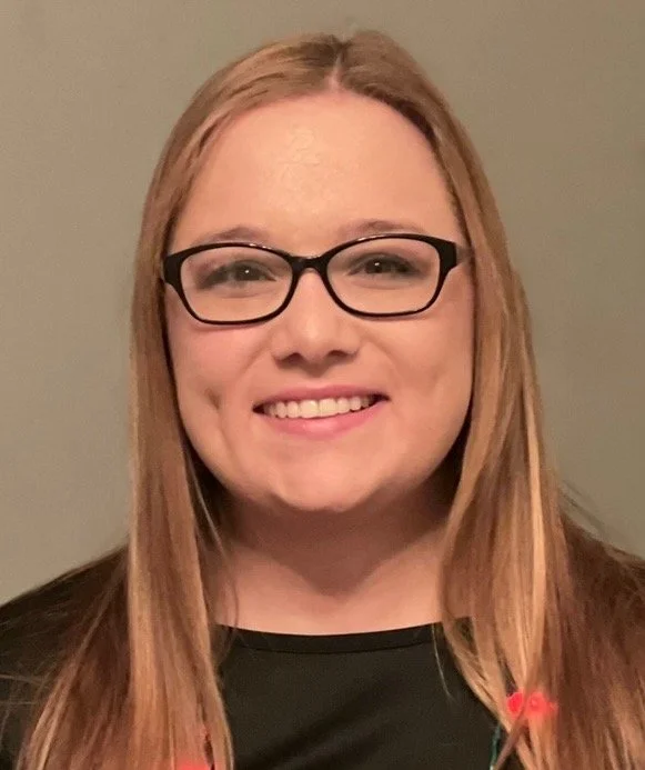 A young woman with long red hair, glasses, and a black shirt, smiling at the camera, in front of a plain beige wall.