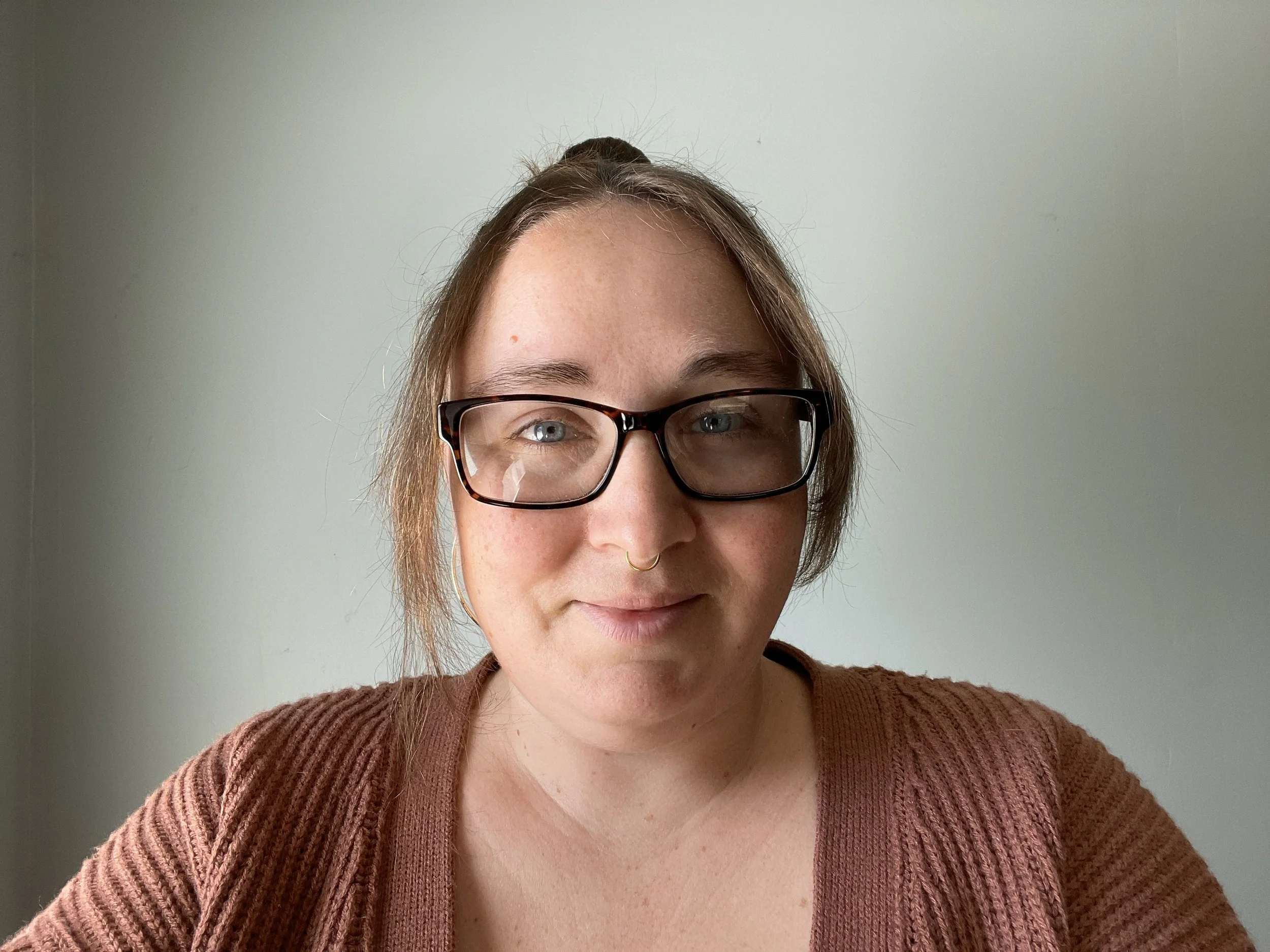 A woman with glasses and a septum piercing taking a selfie against a plain white wall.