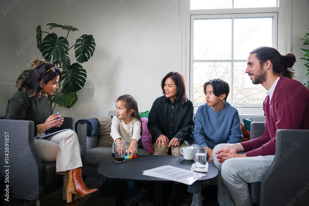Family therapy session in a living room with five people seated on sofas and chairs, engaging in conversation, with a large window and indoor plants in the background.