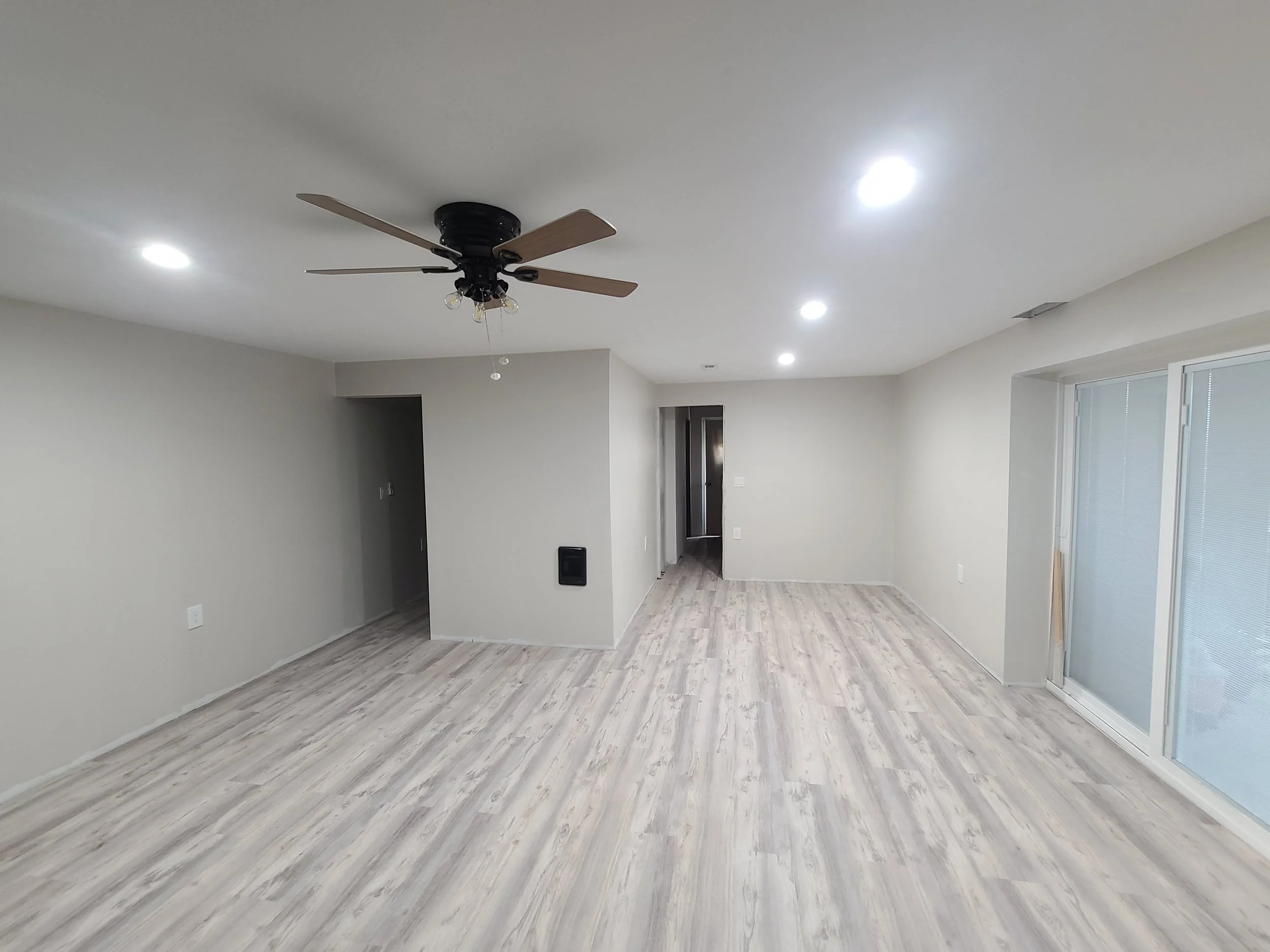 Empty living room with light-colored wooden flooring, white walls, ceiling fan, and sliding glass door.