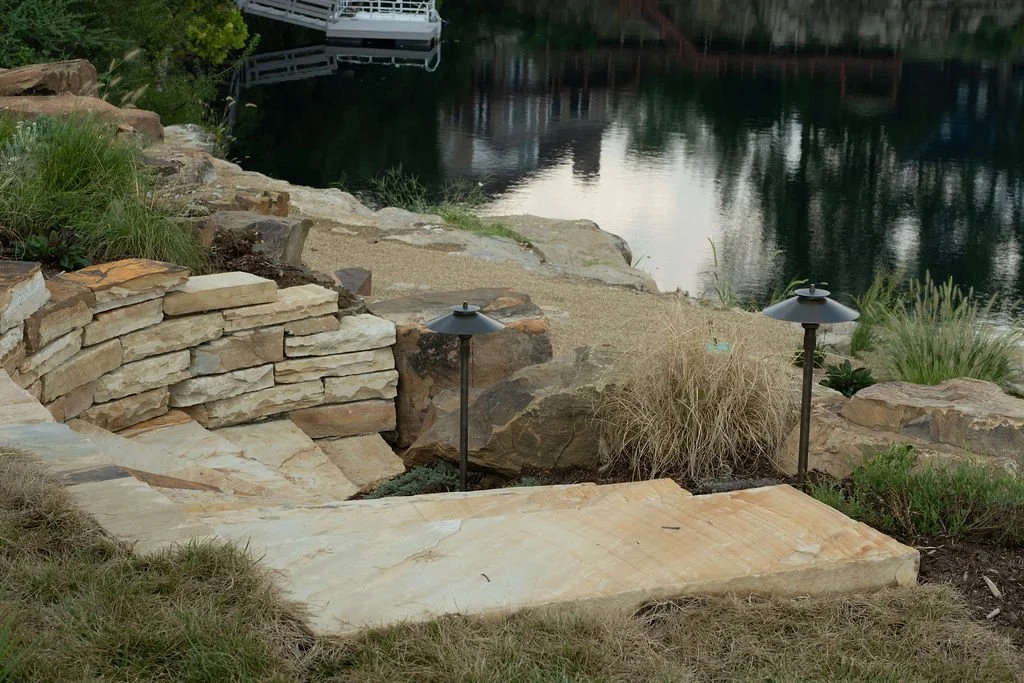 A landscaped area beside a water body with stone steps and rocks, two black outdoor lamps, and various plants and grasses.