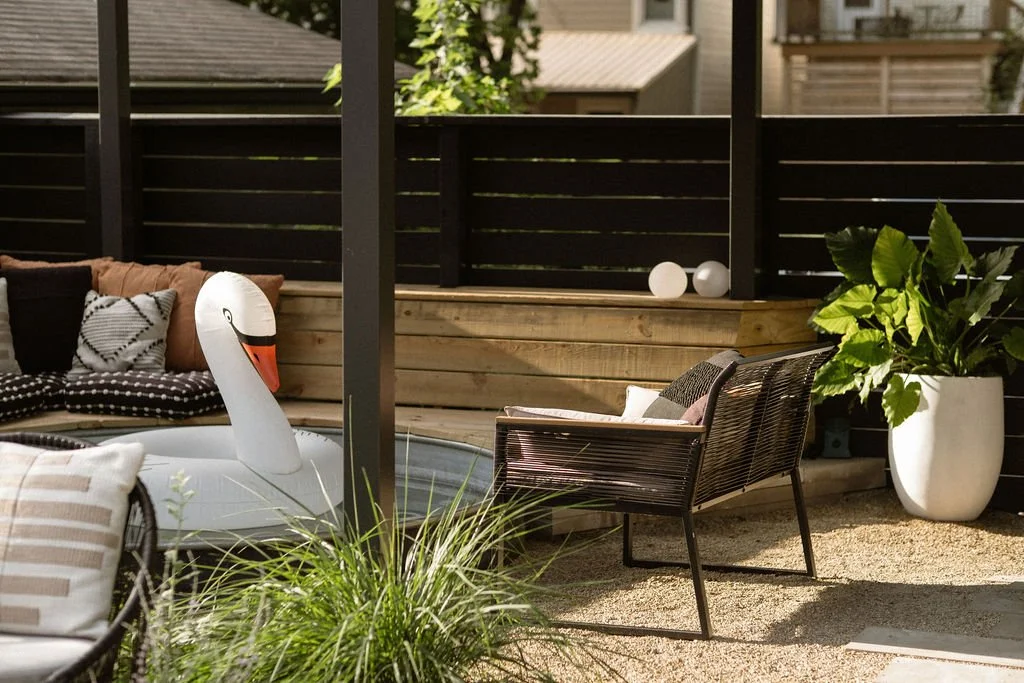 Outdoor patio with black wooden fence, a white inflatable swan pool float, a large potted green plant, a black patio chair with beige cushions, and a wooden bench with pillows.
