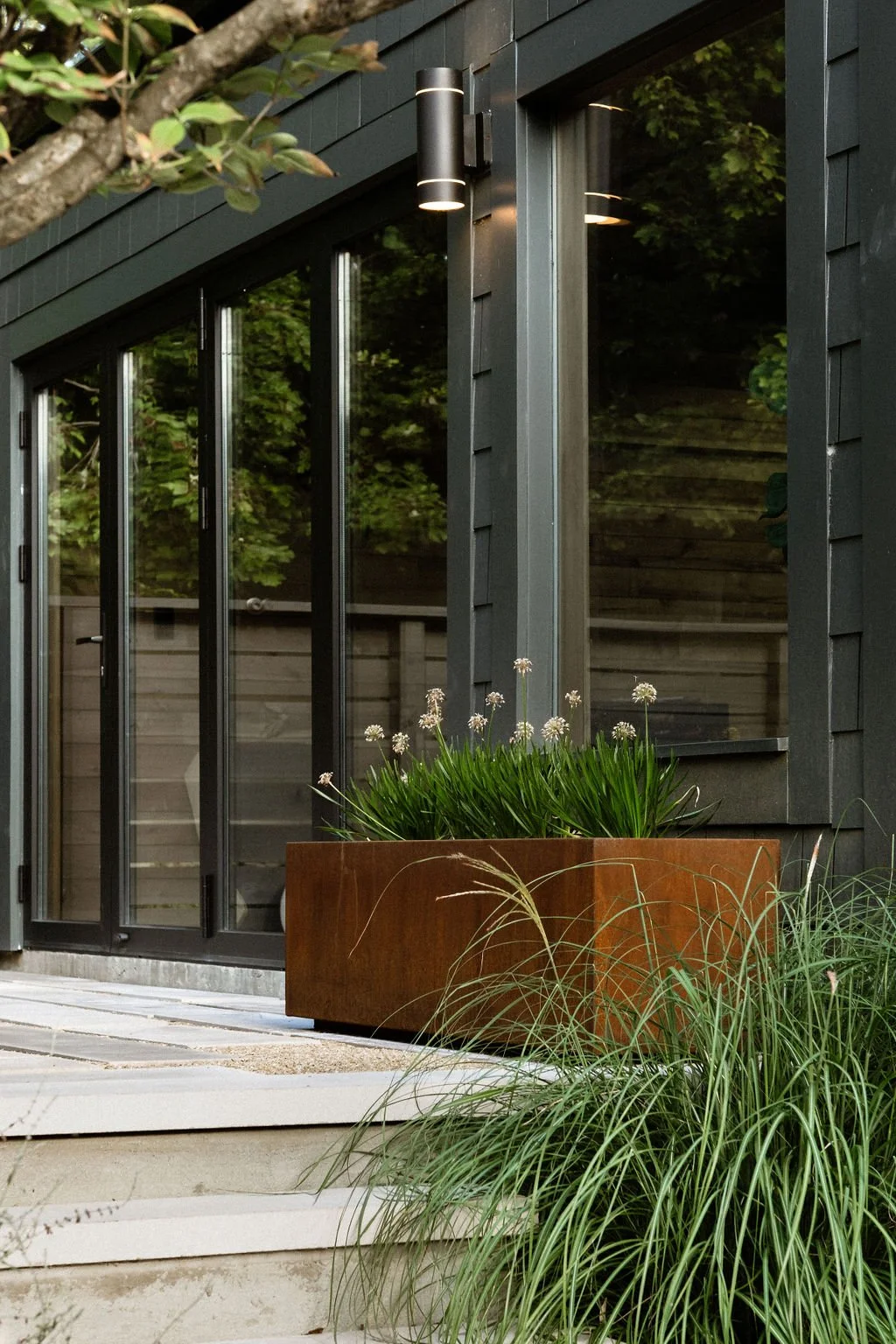 Exterior view of a modern house with large glass sliding doors, a wall-mounted light fixture, and a large rust-colored planter with green plants and white flowers near a wooden deck and steps.