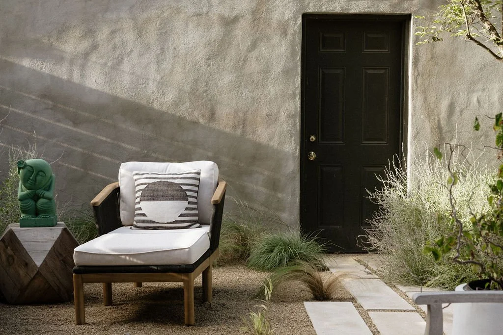 Outdoor patio with a black door, a white cushioned chair with a striped pillow, a small wooden table with a green sculpture, and desert plants.