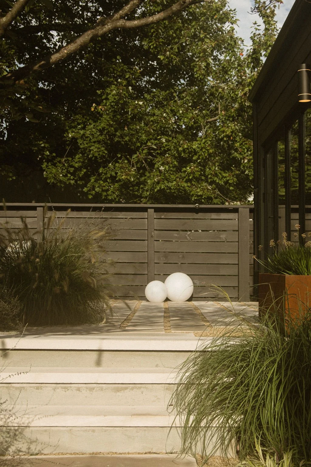 Outdoor patio with a wooden fence, large white decorative spheres, and lush green plants, adjacent to a house with black exterior walls and glass windows, under a leafy tree.