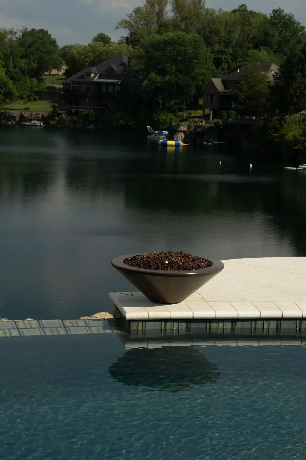 A bowl of dark-colored berries or fruit placed on the edge of a poolside table overlooking a lake with houses and trees.