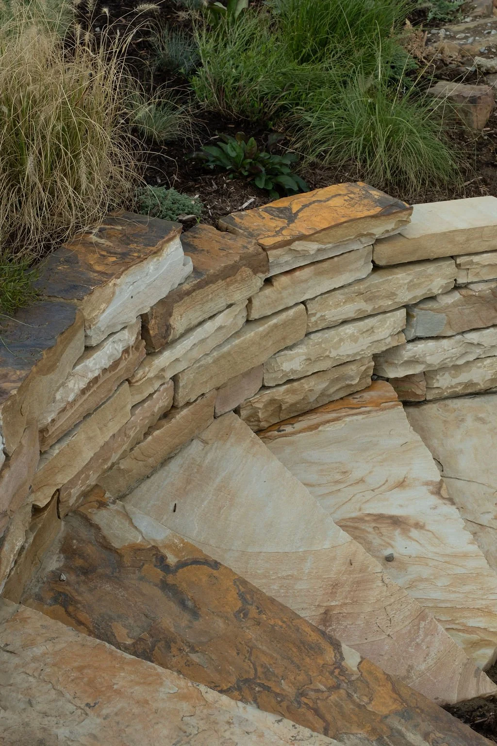 Close-up of stacked flat stones forming a curved stone wall, with some greenery and plants in the background.