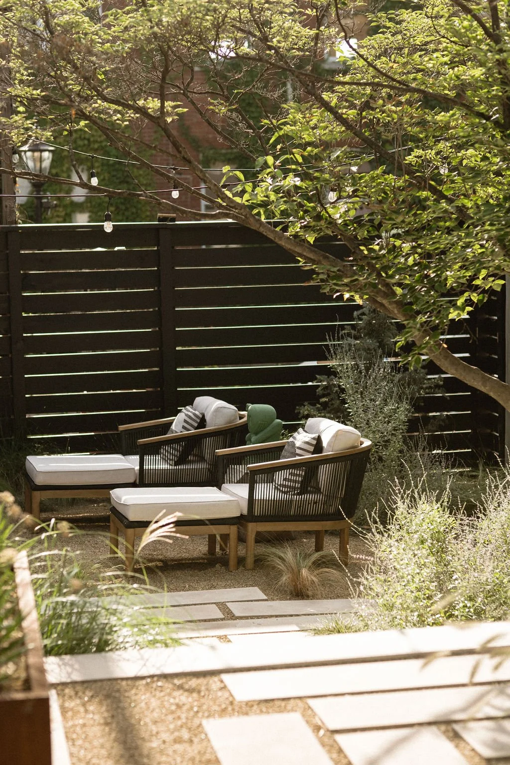 A cozy outdoor patio area with two cushioned lounge chairs and a small green side table, surrounded by plants and shaded by a tree, with string lights hanging above and a black wooden fence in the background.