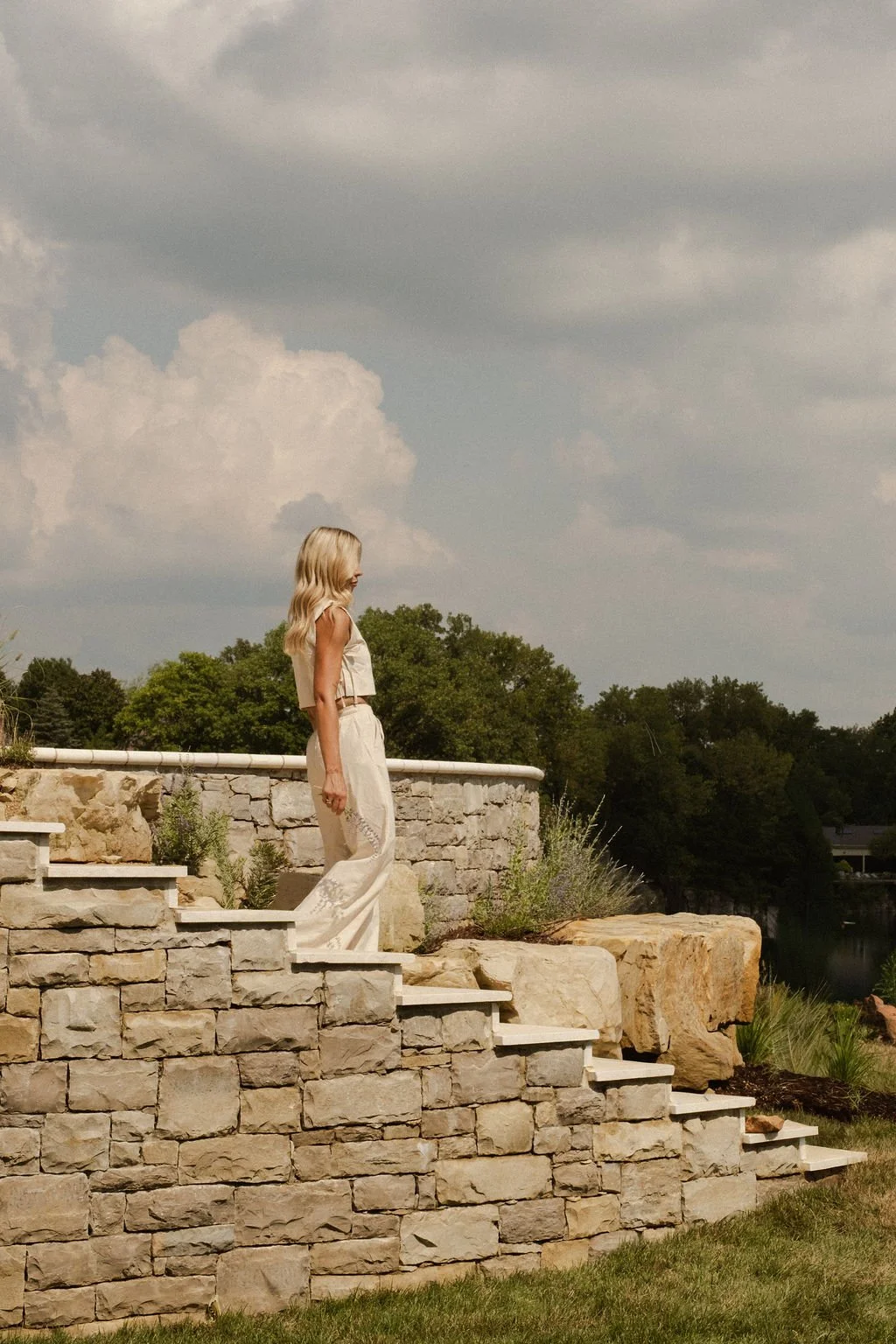 A woman with long blonde hair, wearing a light-colored sleeveless top and matching loose pants, stands on stone steps outdoors, looking to her left. The background features a cloudy sky, green trees, and a stone wall with a railing.