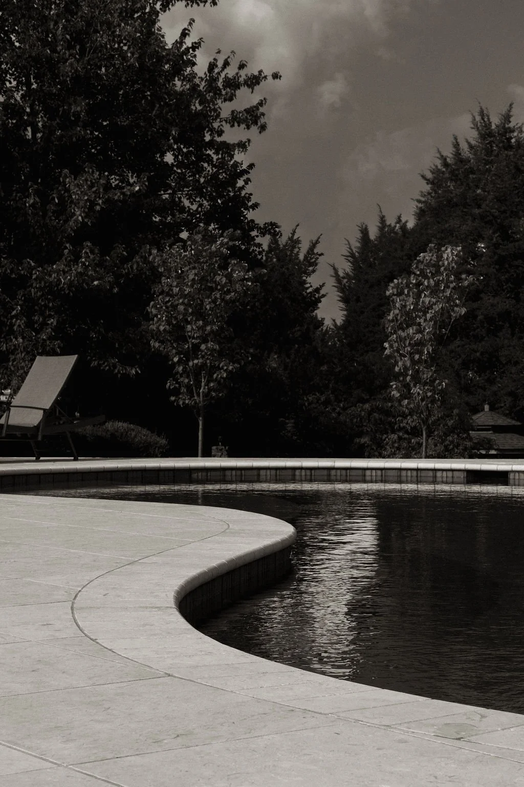 A pool surrounded by a concrete deck with trees and houses in the background on a cloudy night.