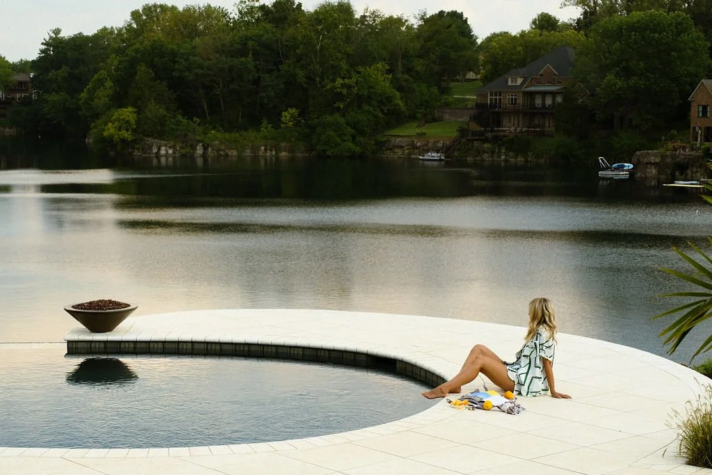 A woman sitting by the edge of an infinity pool overlooking a lake with houses and trees in the background.