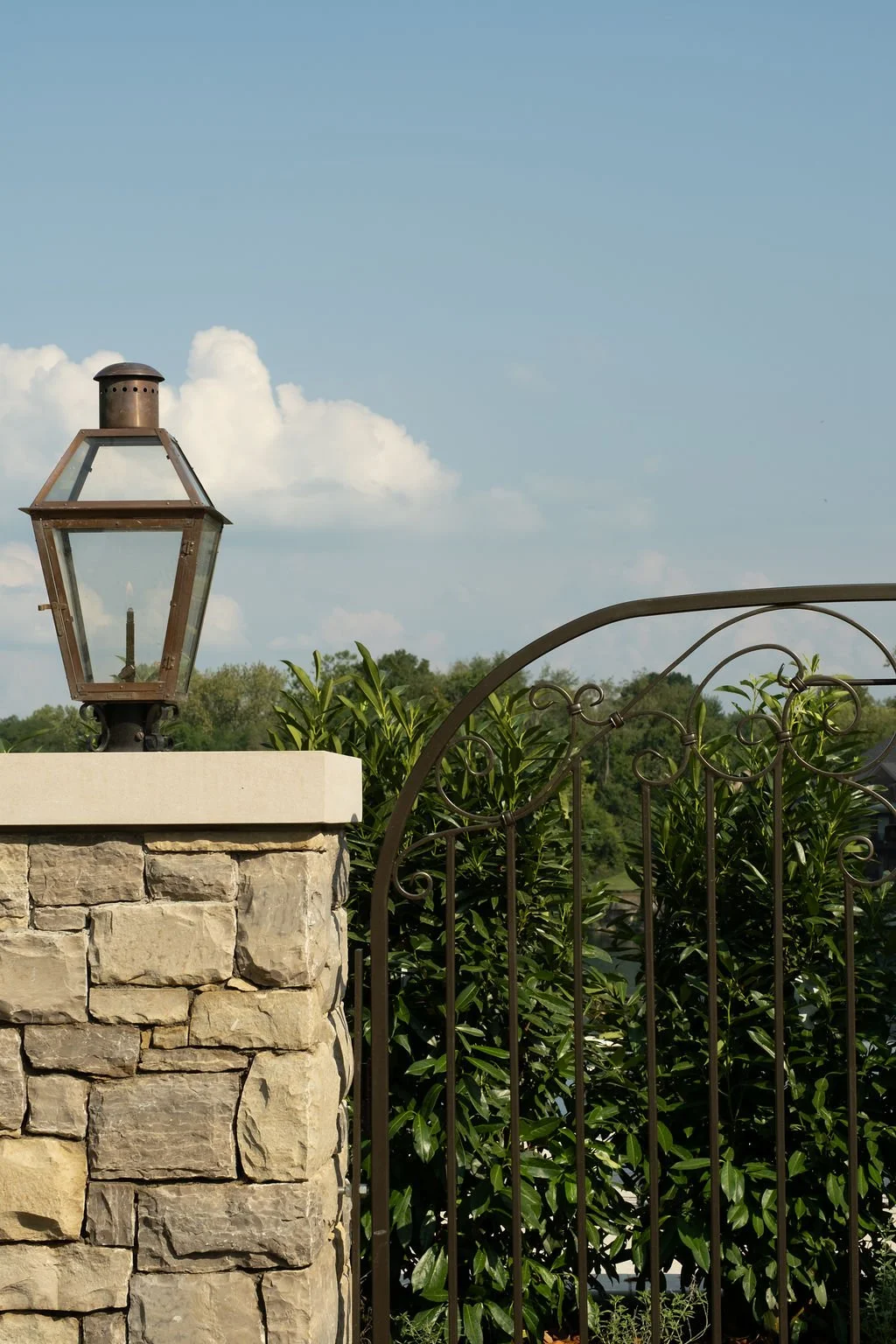 A stone pillar with a black metal outdoor lantern on top, next to a decorative wrought iron gate and lush green bushes, under a partly cloudy sky.