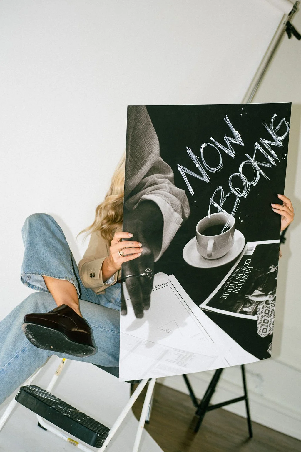 Person holding a large sign with the message "NOW AROUND" and an image of a coffee cup on a black background, partially obscuring their face. The person is seated on a white ladder, wearing jeans, a beige blazer, and black shoes.
