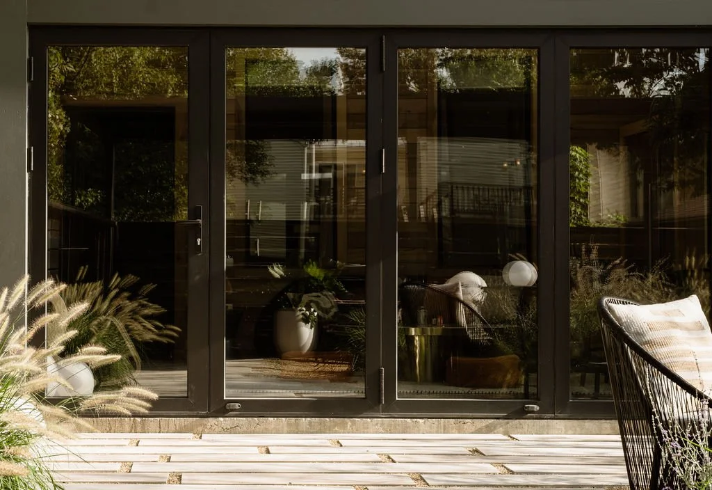 Glass sliding door leading to a patio with outdoor furniture, potted plants, and greenery reflected in the glass.