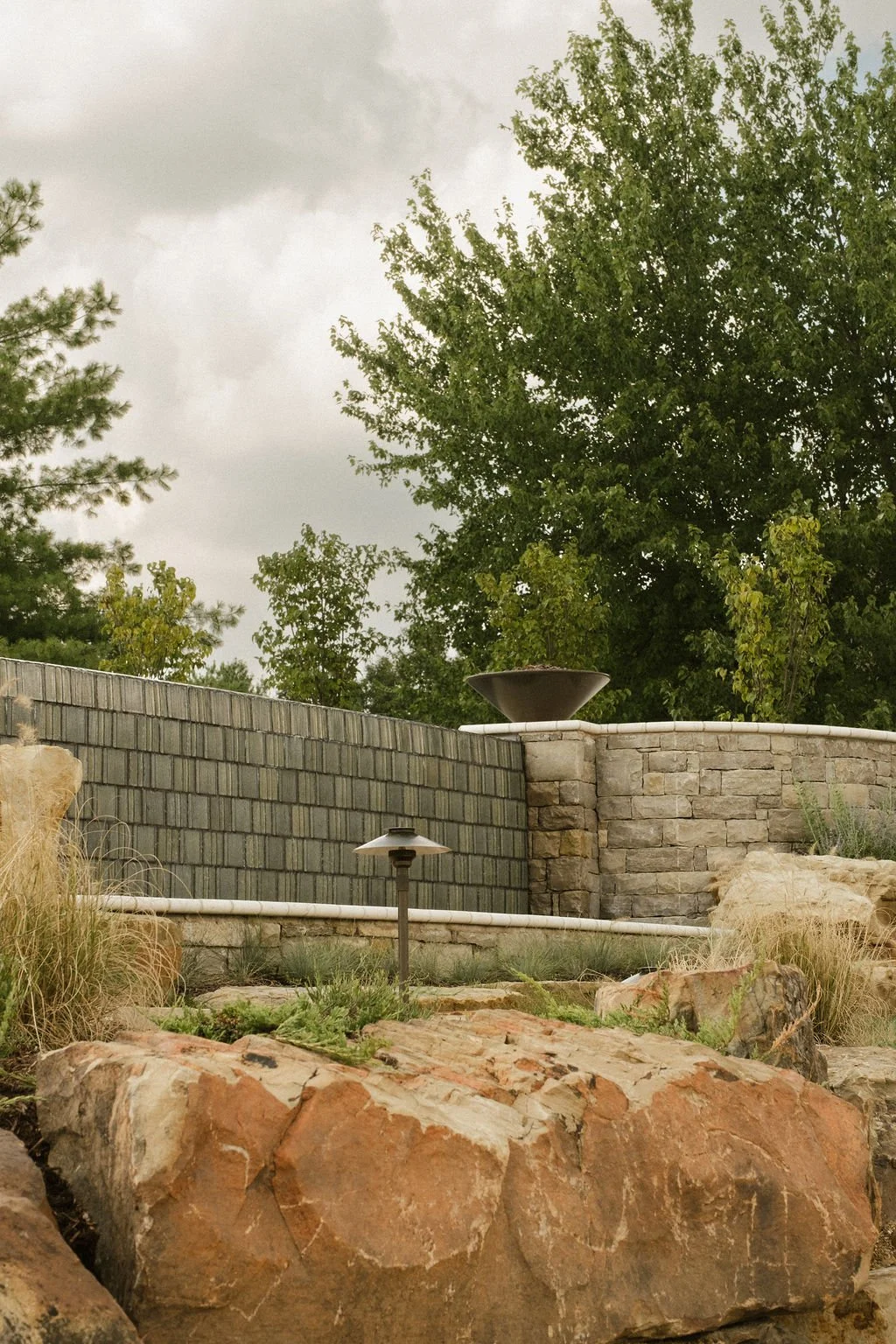 A landscaped backyard with a stone wall, yard lights, large rocks, plants, and tall trees under a cloudy sky.