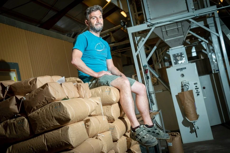 A man in a blue T-shirt and shorts sitting on a stack of wrapped packages inside a warehouse with machinery around.