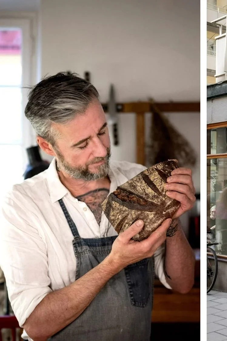 A man with gray hair and a beard wearing a white shirt and black apron holding a loaf of artisanal bread in a home kitchen.