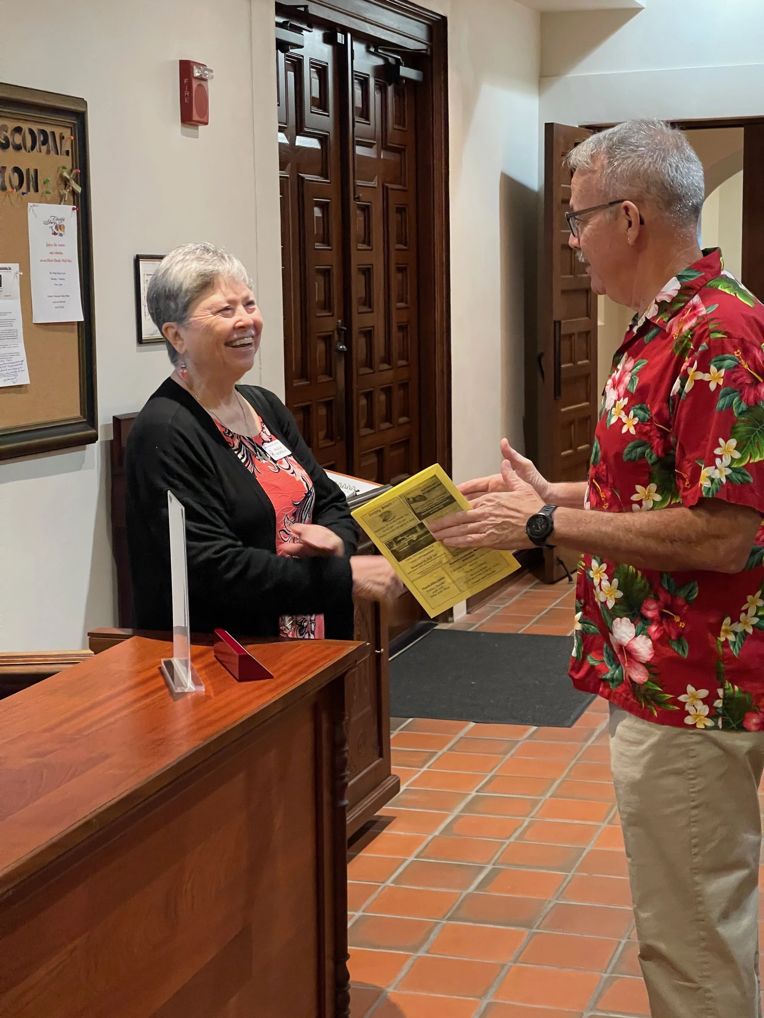 A woman with short gray hair and a black blazer is smiling and talking to a man with gray hair, glasses, wearing a red Hawaiian shirt with yellow, green, and white flowers. They are standing in a reception area, and the woman is behind a wooden counter. The man is holding yellow pamphlets and appears to be engaged in conversation.