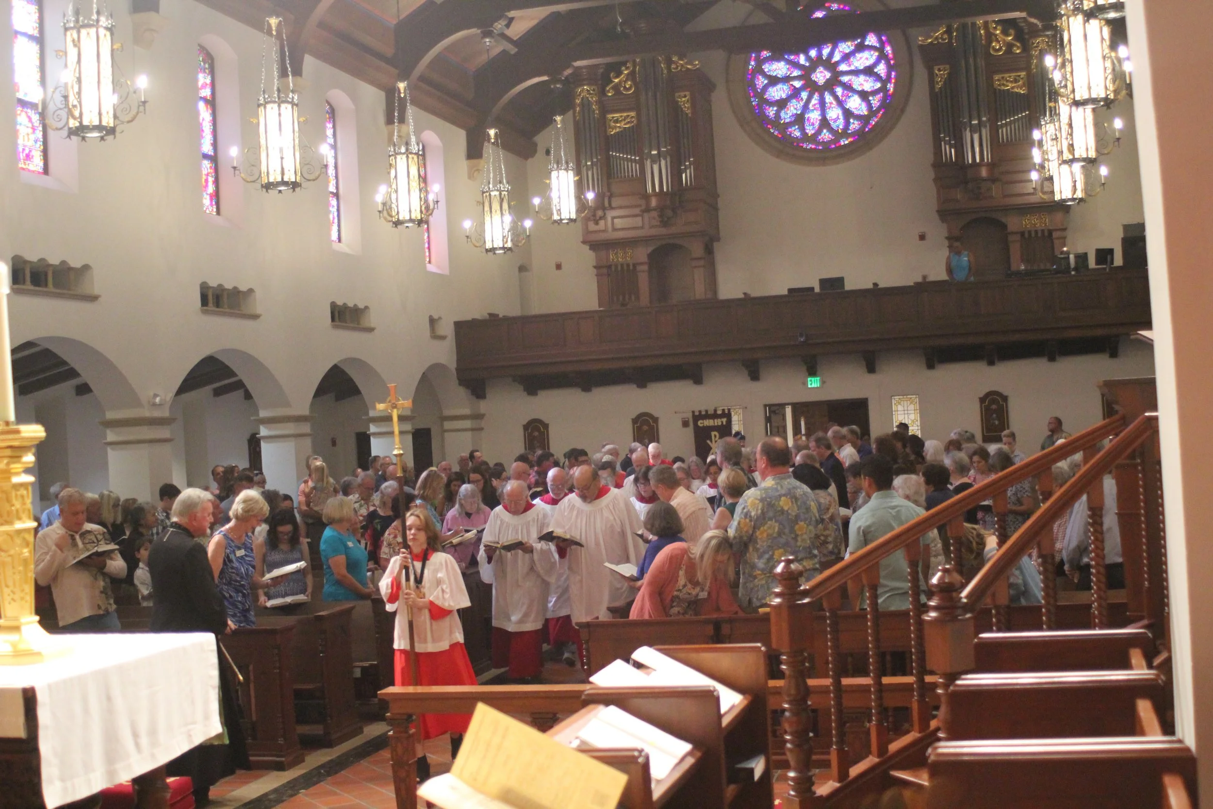 A church interior filled with people participating in a religious service, with stained glass windows, wooden pews, and clergy dressed in white robes.