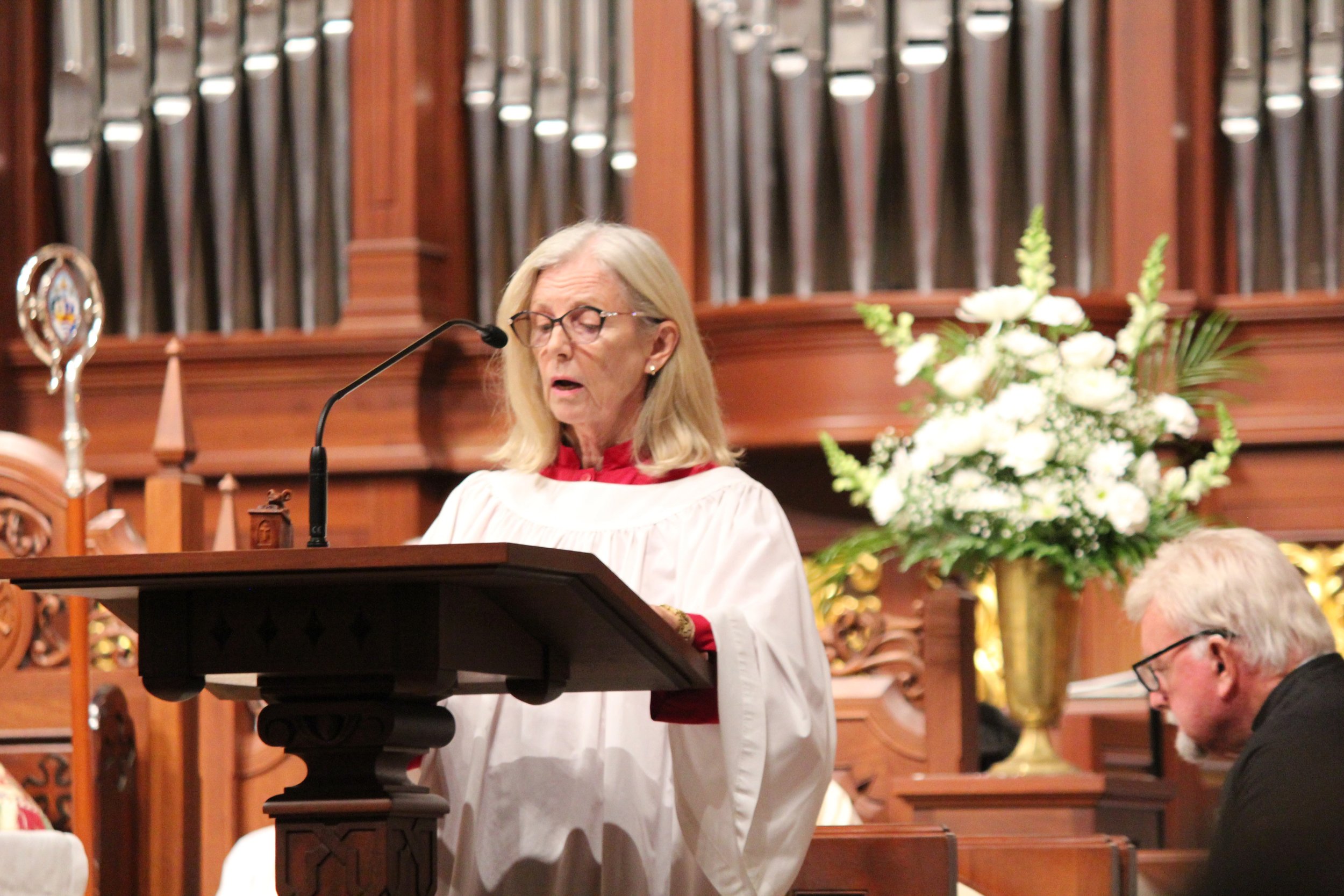 A woman dressed in white robes stands at a pulpit reading during a religious service inside a church. A man with glasses, gray hair, and a beard is seated to her right, looking downward. There is a large bouquet of white flowers in a gold vase to the right of the woman, and a pipe organ in the background.