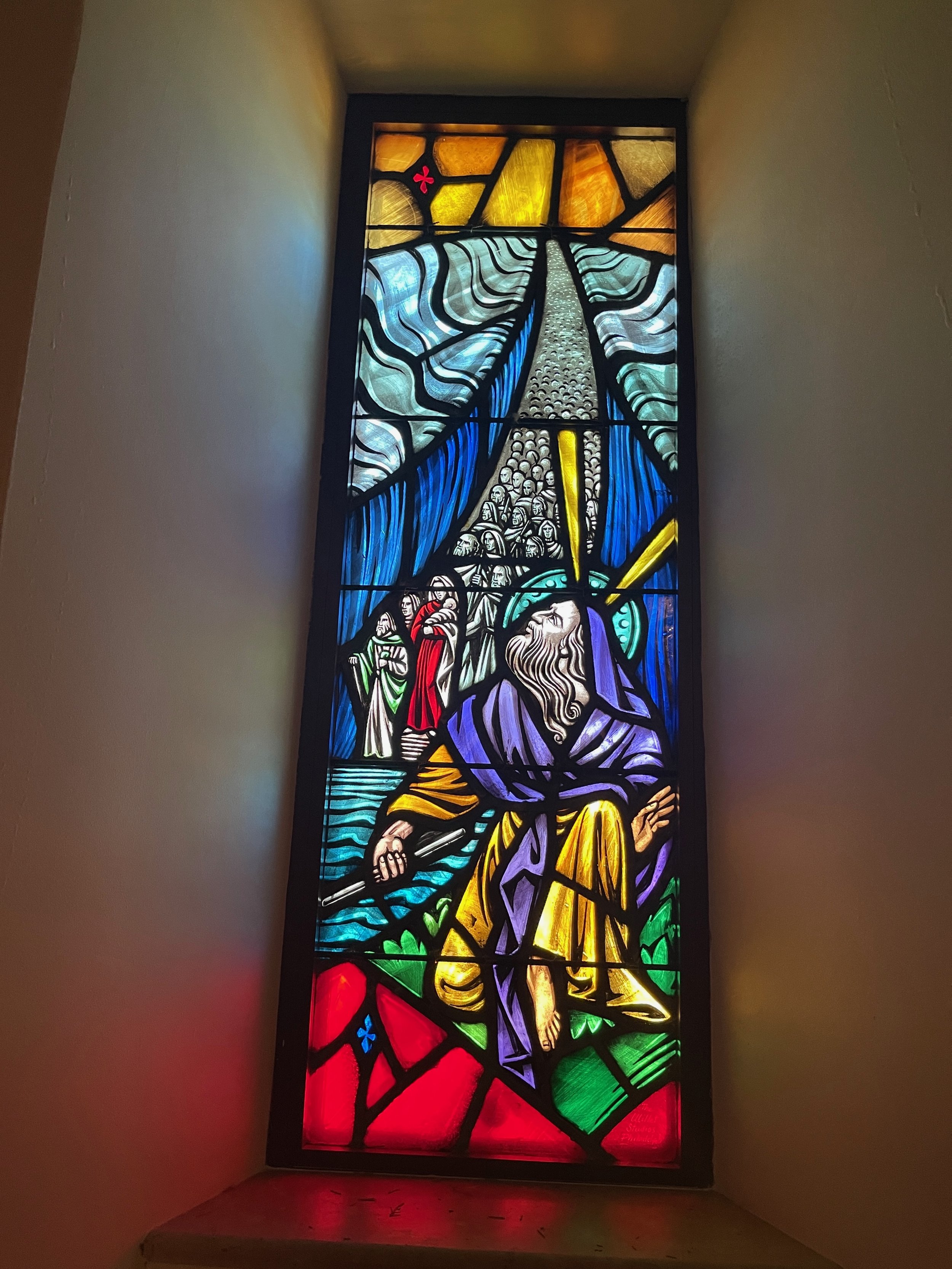 Colorful stained glass window depicting a biblical scene with a bearded man in purple and yellow robes kneeling or praying, a group of people in the background, and divine rays or light shining down from above.