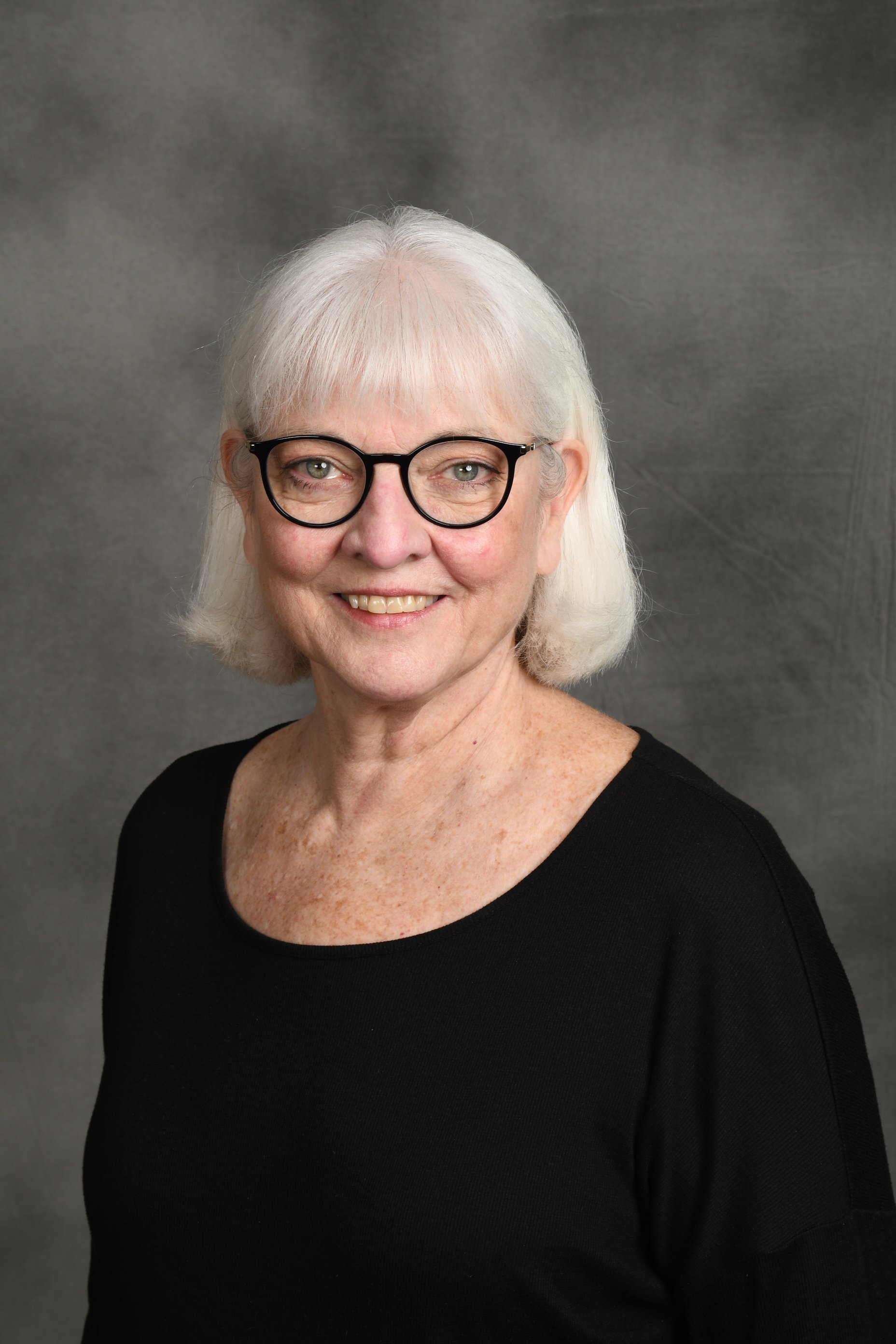An elderly woman with short white hair, wearing glasses and a gray t-shirt, smiling with arms crossed against a neutral gray background.