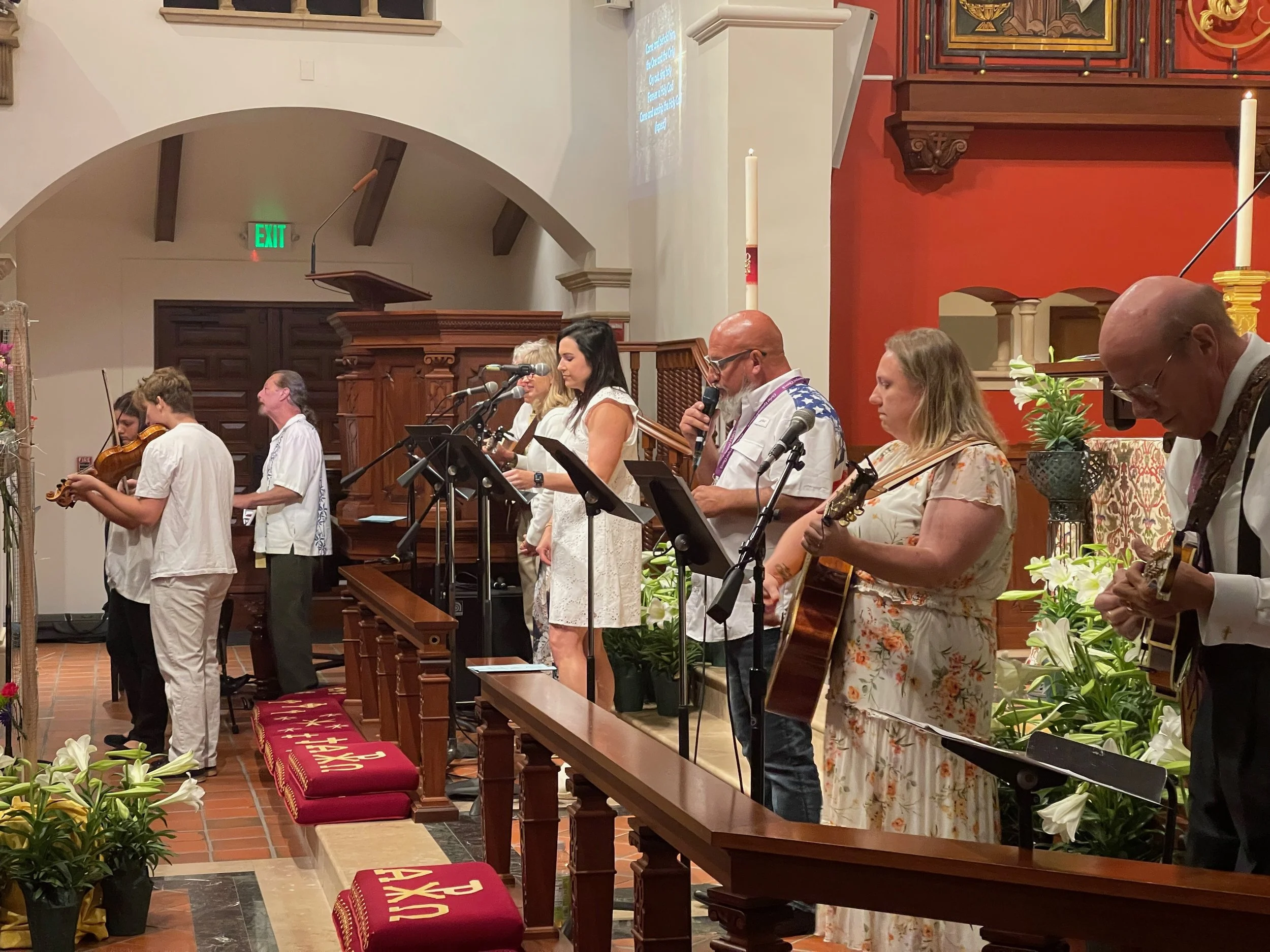 Group of people singing and playing musical instruments on a stage inside a church or a chapel, with flowers and candles nearby.
