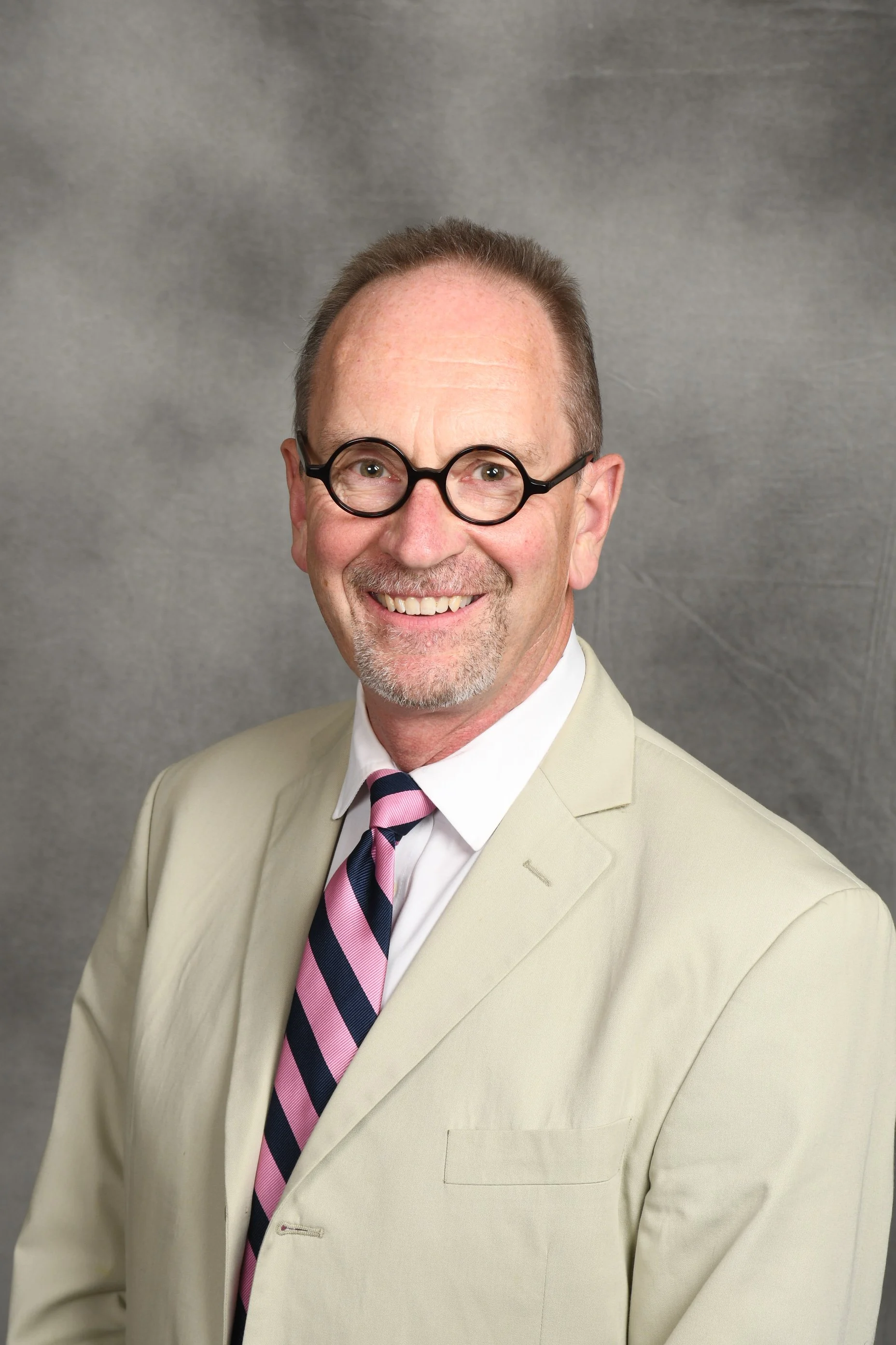 A man with glasses, wearing a dark suit and red tie, sitting and smiling indoors with warm lighting and paintings on the wall behind him.