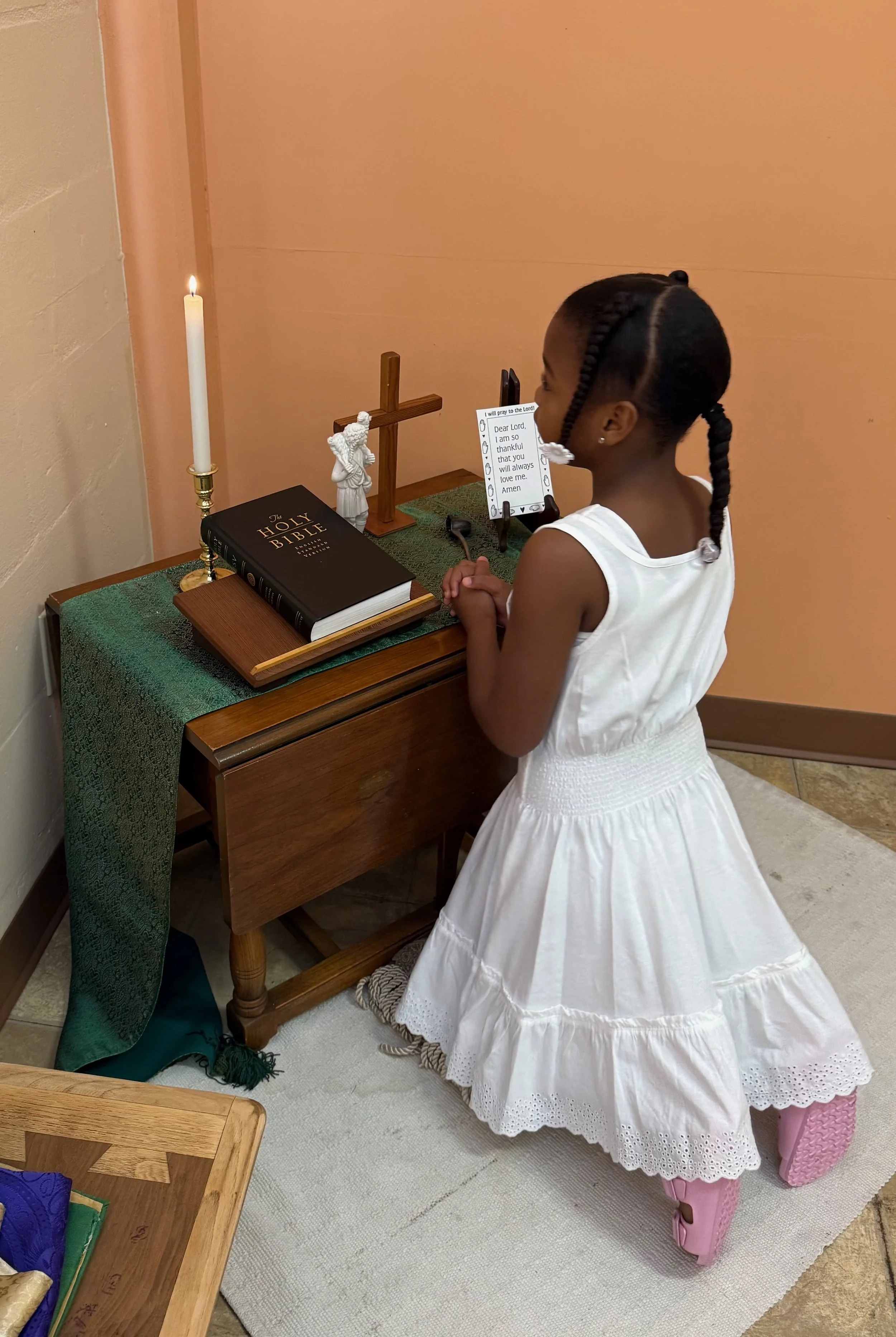 A young girl in a white dress kneels in prayer at a small wooden altar with a Bible, a cross, a statue of Jesus, a lit candle, and a tablet displaying a prayer. She is facing the altar with her hands clasped.
