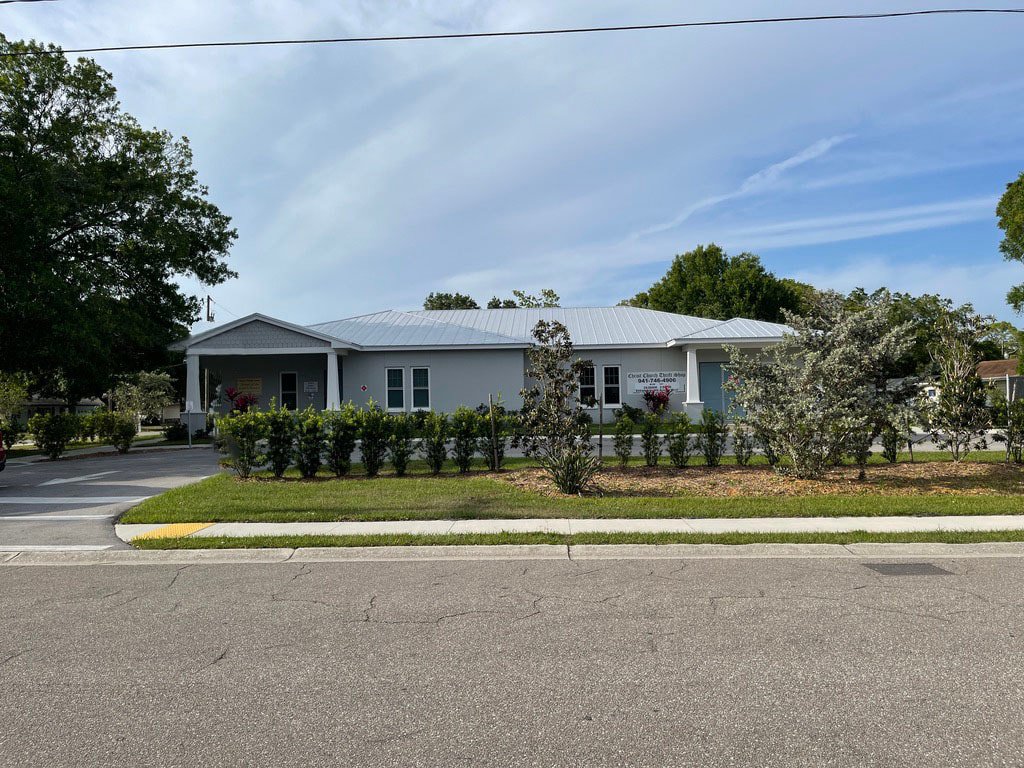 A light blue single-story building with a metal roof, surrounded by bushes and trees, with a parking lot in front.