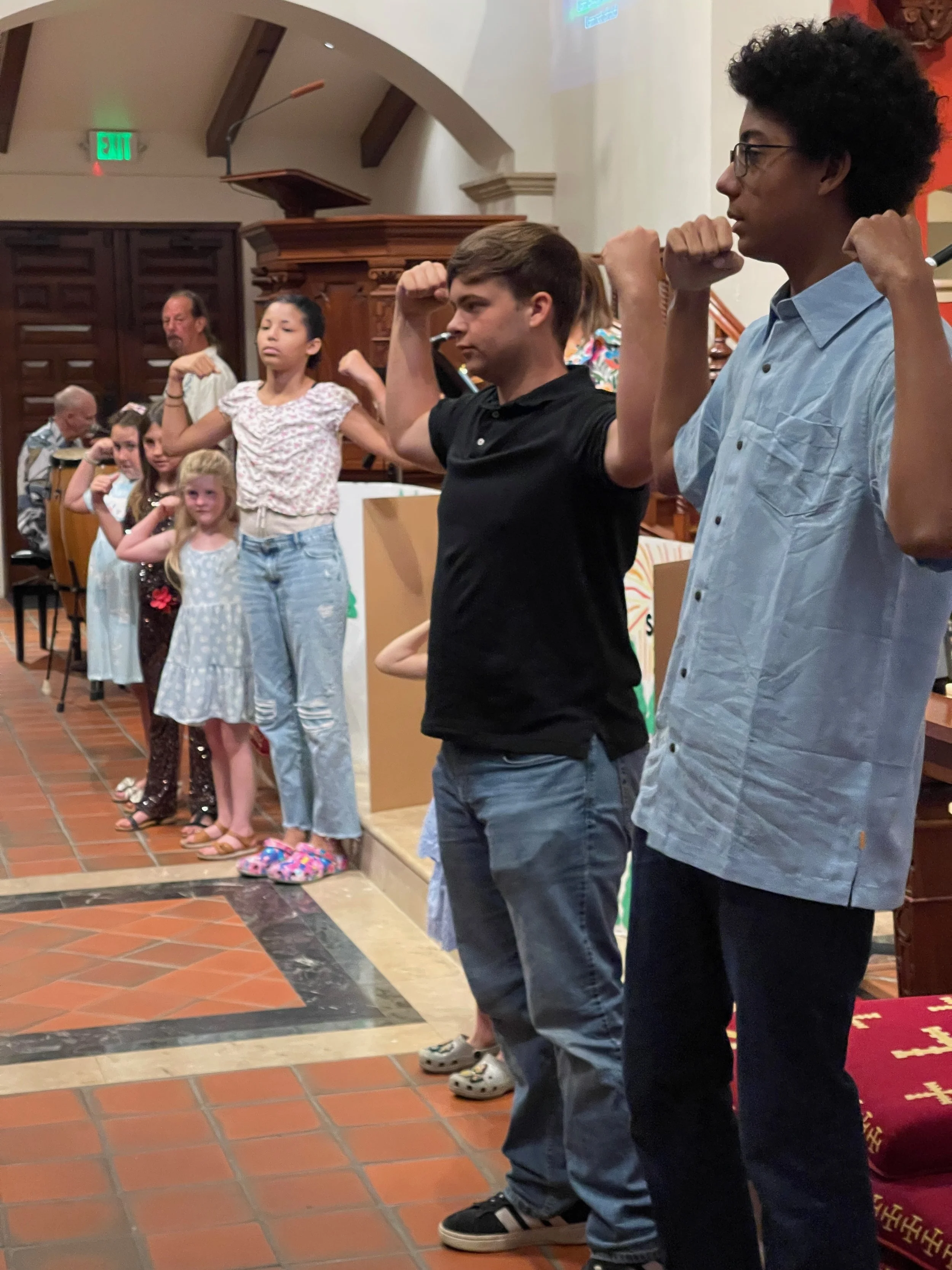 A group of children and teenagers standing in a row, raising their arms in a flexing pose inside a church or similar venue.