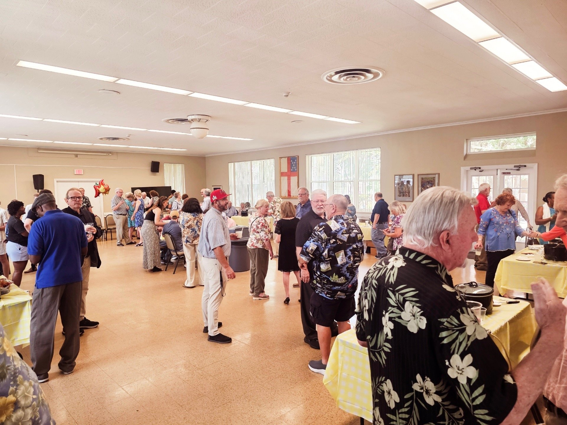 People standing in line and socializing at a buffet-style gathering in a community center or hall with yellow tablecloths, large windows, and framed artwork on the walls.