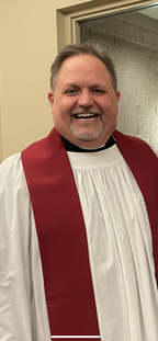 A man dressed in a white choir robe with a red stole, smiling indoors.