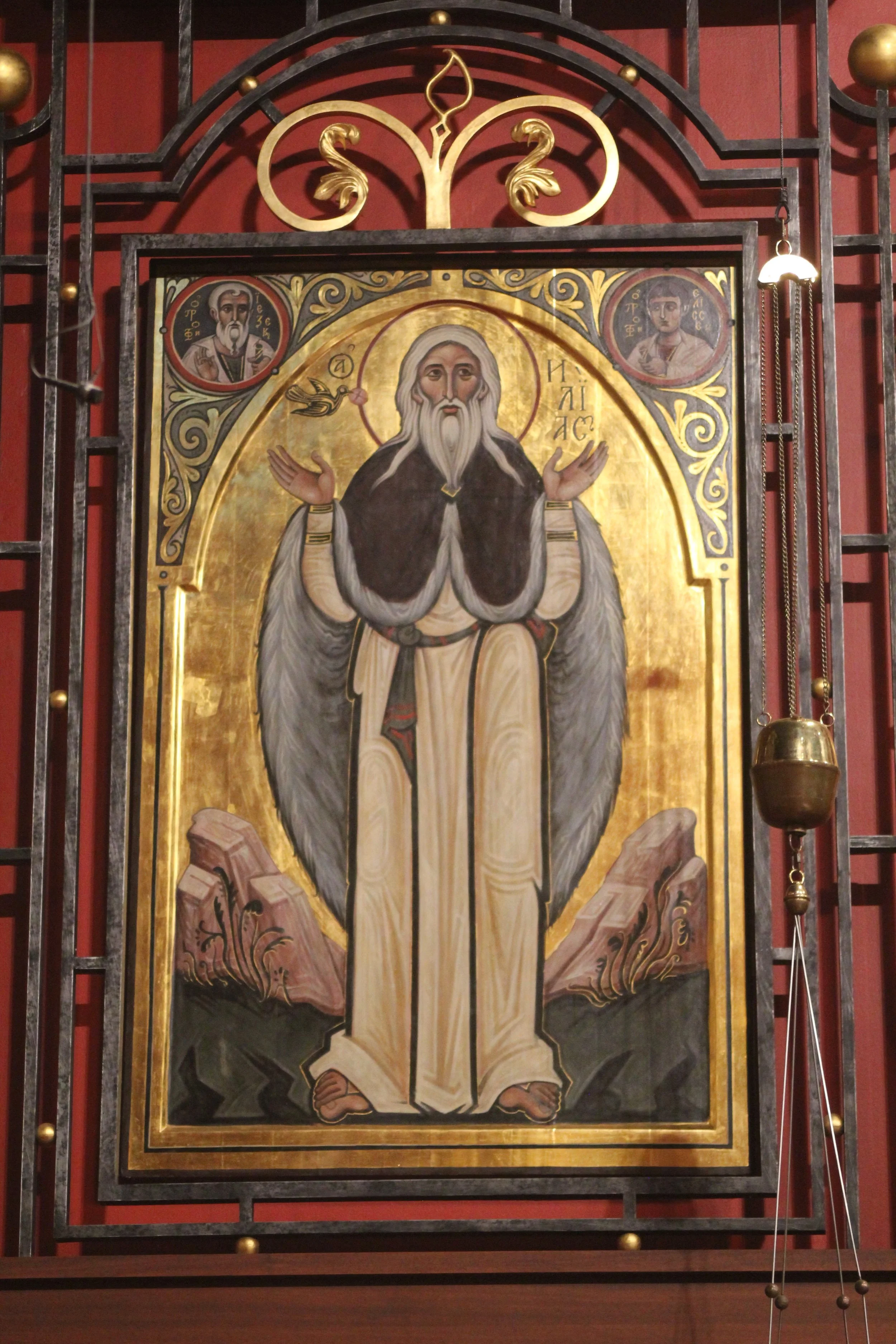 Religious icon painting of Jesus Christ with arms raised, surrounded by gold background, with two smaller portraits of saints in circles at the top corners, set within a metal frame in a church interior.