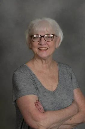 An elderly woman with short white hair, wearing glasses and a gray t-shirt, smiling with arms crossed against a neutral gray background.