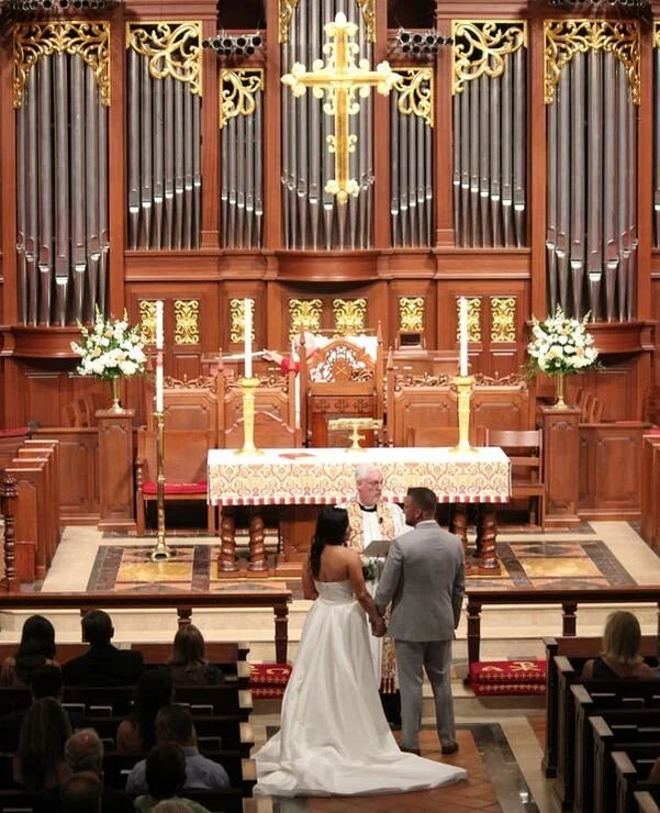 A couple getting married at the front of a church with a priest, with an ornate wooden altar and large organ pipes behind them. They are standing before an officiant, holding hands, with guests seated in pews watching.