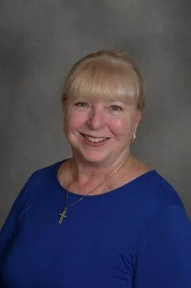 A portrait of a smiling woman with blonde hair and bangs, wearing a blue top and a cross necklace, against a gray background.