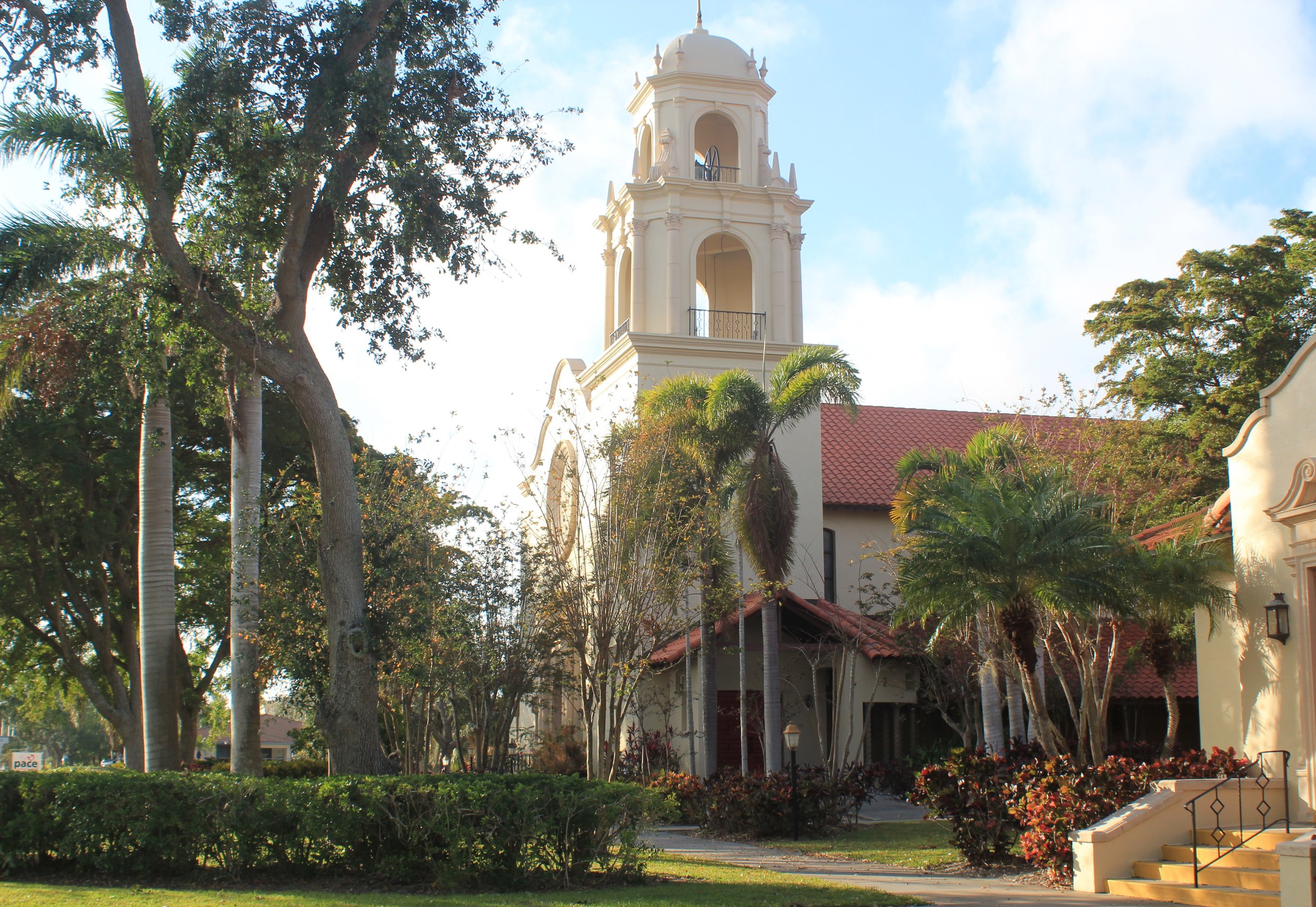 A church with a tall white bell tower surrounded by green trees and shrubs, with a clear sky background.