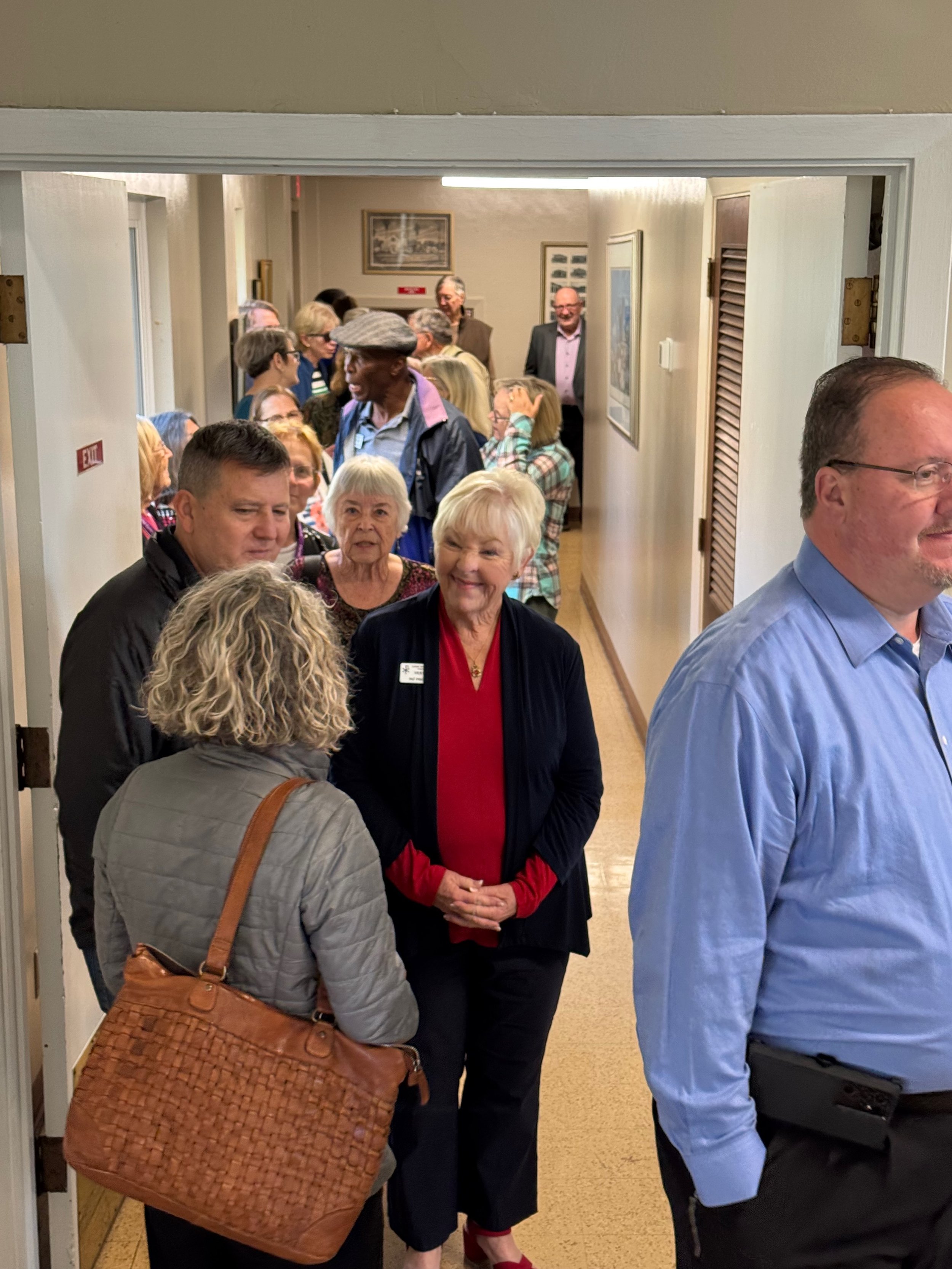 People standing and talking in a hallway, some in line, with painting on the wall and an exit sign visible.