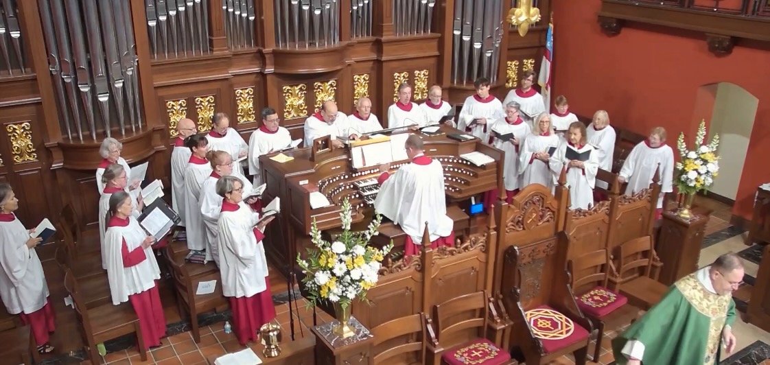 A choir of people dressed in white robes with red accents singing in a church, with an organ and floral arrangements nearby.
