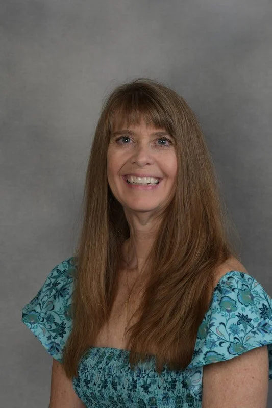 A woman with long brown hair wearing a blue floral dress, smiling against a gray backdrop.
