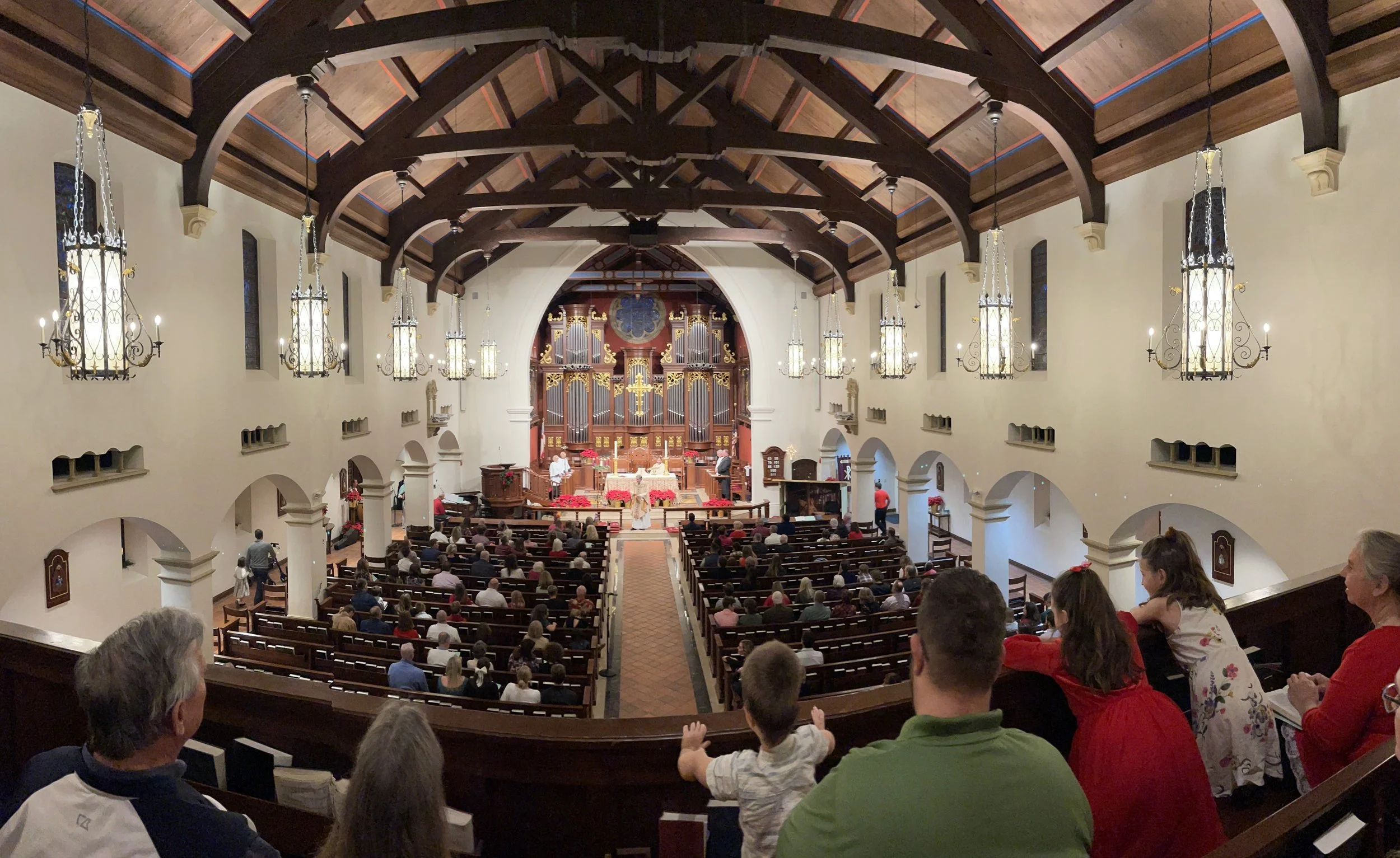 Interior view of a church filled with people attending a service, with an ornate pipe organ in the background and chandeliers hanging from the wooden vaulted ceiling.